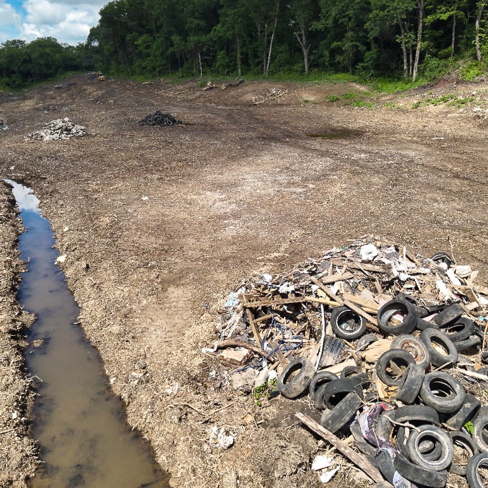 Trash and vegeation cleanup site with scattered debris and tires near a muddy drainage channel.