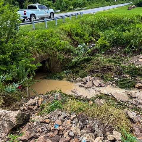 A work truck is parked on a street above an embankment that has collapsed due to extreme flooding. Water is flowing into an aged culvert and damage is apparent.
