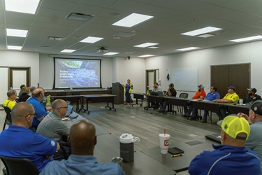 A photograph of people in a training room sitting around a large table looking at a presentation screen.