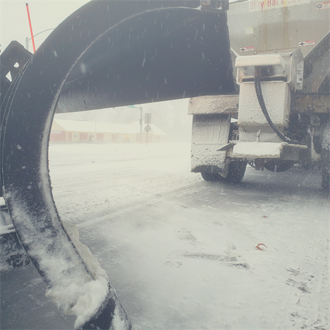 A photograph of a snowplow during a storm with a salt spreader in the background